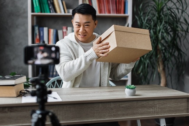 Handsome asian man in casual wear recording video on camera while unpacking gift box. Male blogger sharing his emotions with his subscribers in social networks.